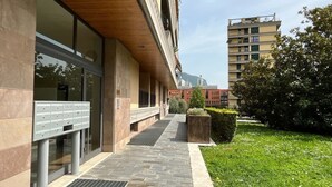 A modern building with a stone facade, a metal mailbox, and a paved walkway leading to a grassy area with a tree and a bush. - La Piazzetta Verde - Affitti Brevi Italia (Lecco)