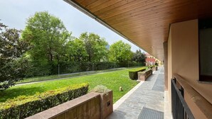 A covered outdoor area with a view of a well-maintained garden and a building in the background. - La Piazzetta Verde - Affitti Brevi Italia (Lecco)