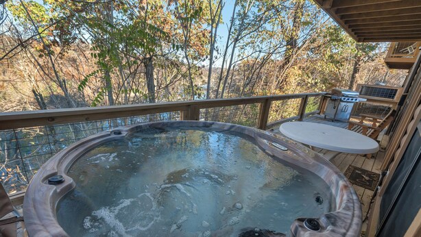 Un jacuzzi en una terraza de madera con vistas a árboles y una zona de barbacoa.