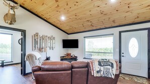 A cabin interior with a wooden ceiling, a brown leather sofa, a mounted deer head, a flat-screen TV, and a welcome sign on the door. - Arkansas Retreat w/ Deck, Fire Pit & Mtn Views! (Jasper)