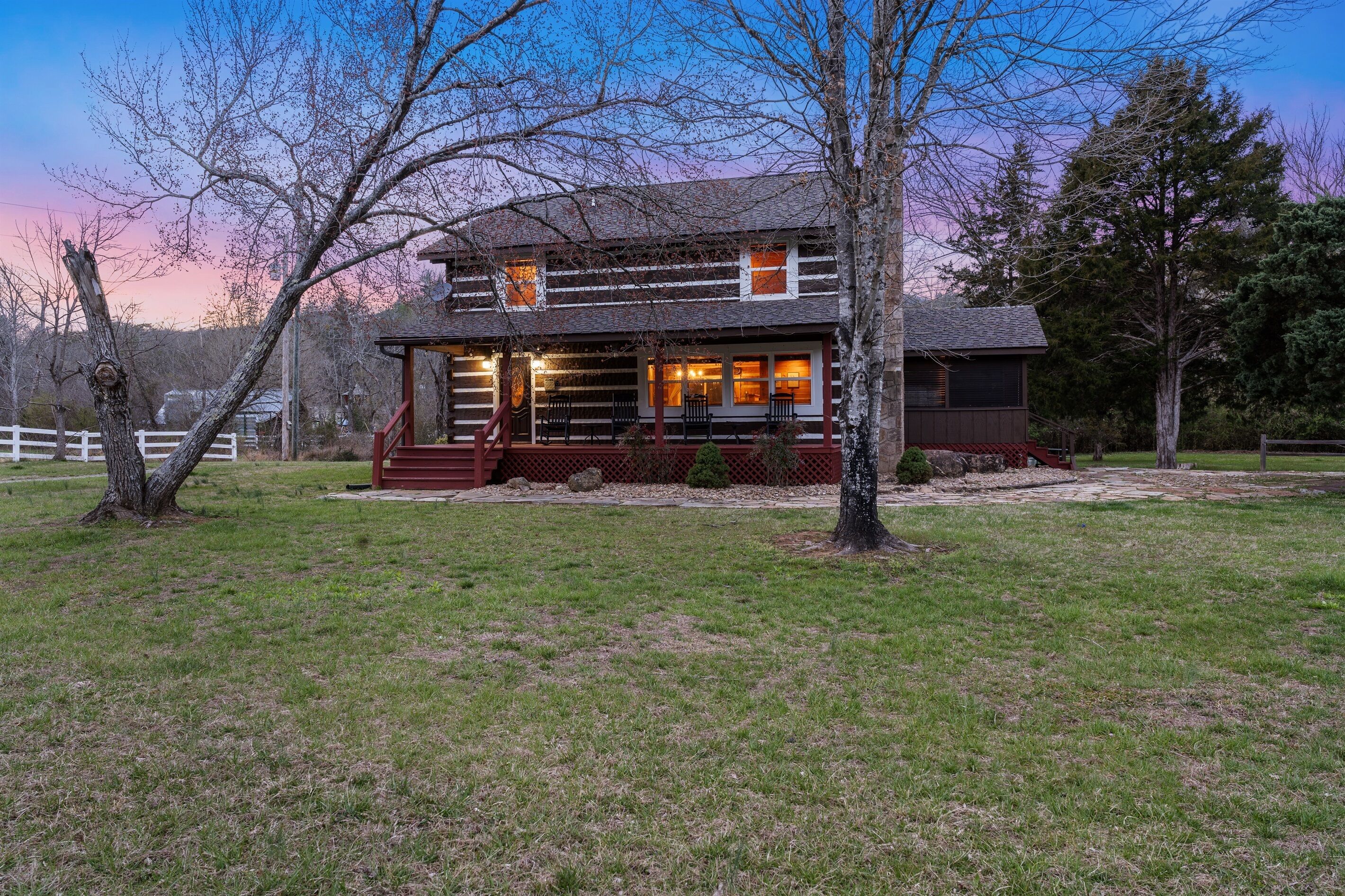 A two-story log cabin with a porch, surrounded by a fenced yard and trees.