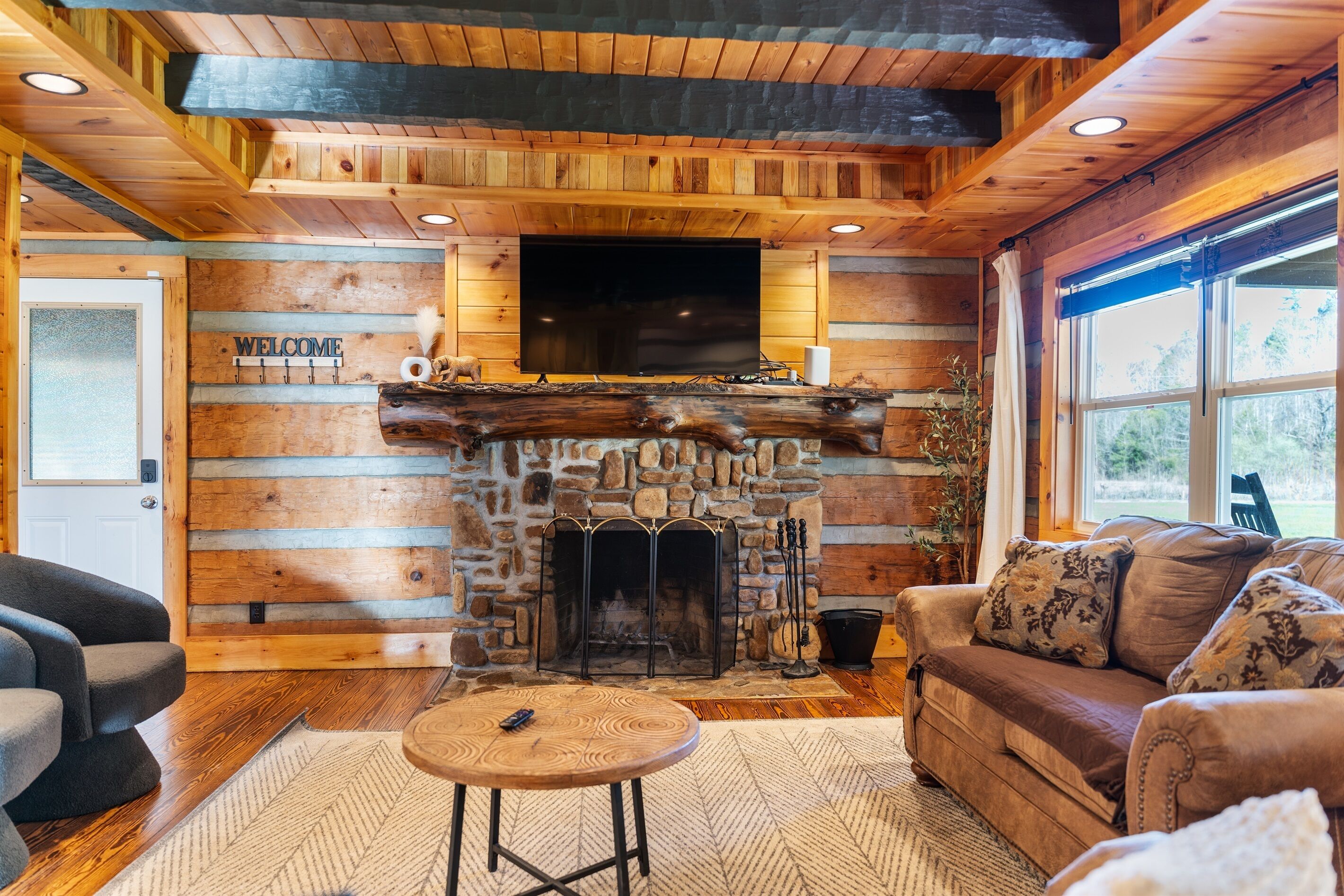 A log cabin interior with a stone fireplace, wooden walls, a flat-screen TV, and a leather sofa.