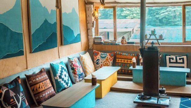 A cozy room with a wooden bench, colorful cushions, a large window with a view of greenery, and a rustic stove. - Young Monk Hostel & Cafe Old Manali (Manali)