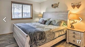 Een slaapkamer met een houten bed, een nachtkastje met een lamp, een raam met zonwering en een wanddecoratie.