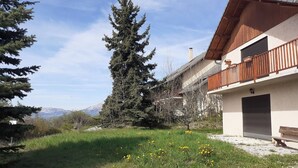Une maison avec un balcon, un garage et une cour verdoyante agrémentée d’arbres et de fleurs.