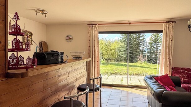 Un salon comprenant un canapé, une table basse et un coin repas avec un bar. Une grande fenêtre, ornée de rideaux, offre une vue sur un jardin.