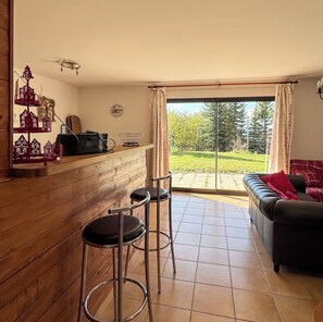 A living room with a sofa, a coffee table, and a dining area with a bar. There is a large window with curtains, and a view of a garden outside.