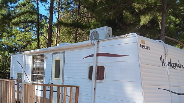 A stationary RV with a wooden deck and a skylight, parked in a wooded area.