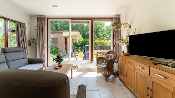 A spacious living room with a sofa, a television, and a wooden cabinet. There is a sliding glass door leading to an outdoor area with a table and chairs. - Holiday Home Zeeland Near Westerschelde (Baarland)