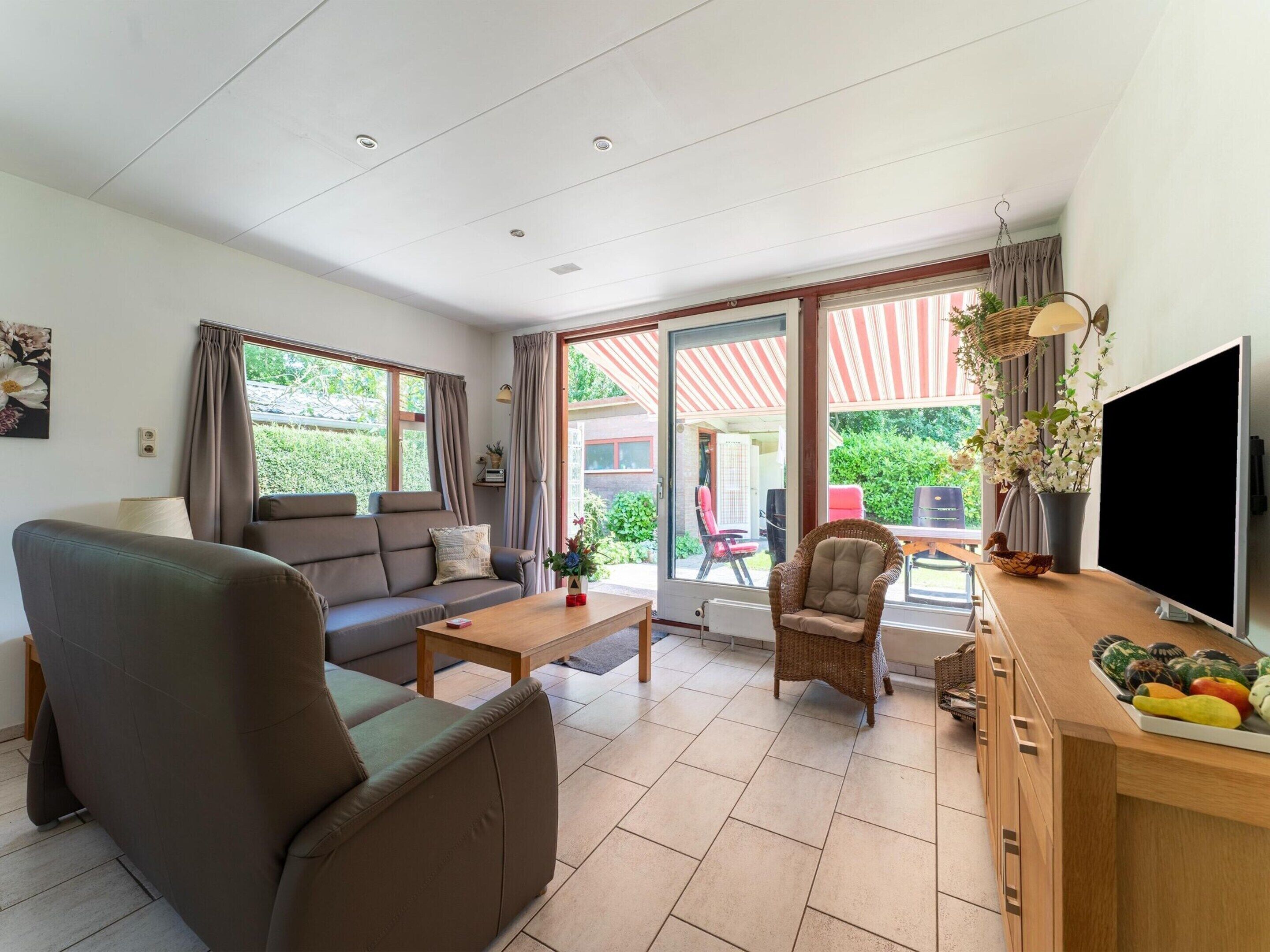 Een moderne woonkamer met een bank, fauteuil en salontafel. Er staat een televisie op een houten dressoir en een raam met gordijnen.