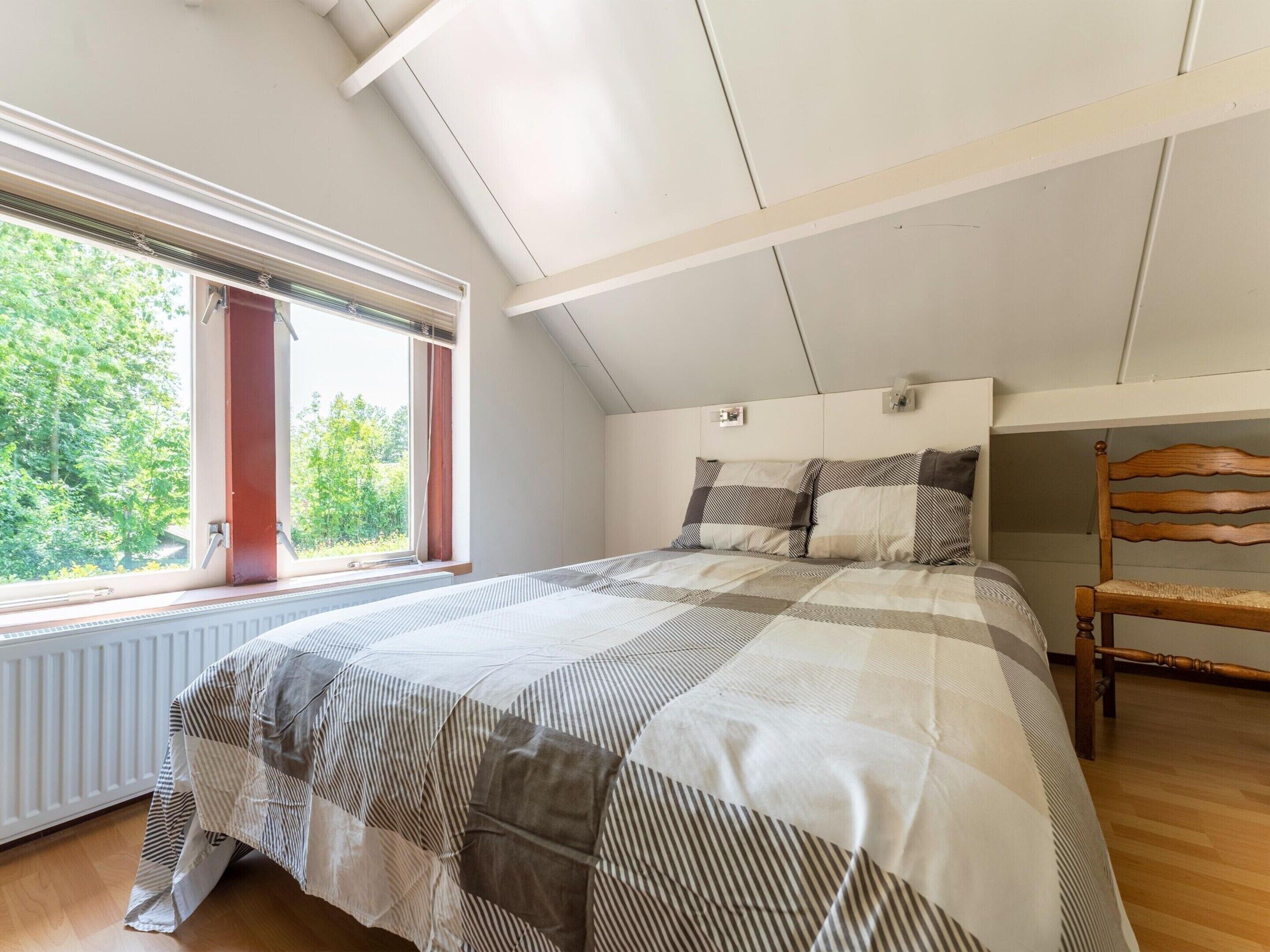 Un dormitorio con techo inclinado, un ventanal grande, una cama con ropa de cama a rayas y una silla de madera.