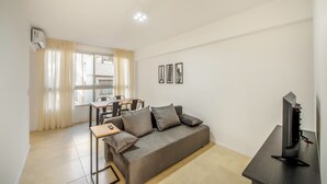 Ein modernes Wohnzimmer mit grauem Sofa, einem kleinen Tisch, einem Fernseher auf einem Lowboard und einem Essbereich mit Tisch und Stühlen.