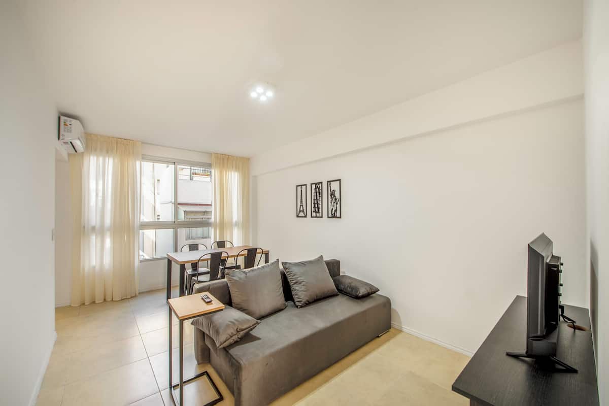 A modern living room with a grey sofa, a small table, a TV on a stand, and a dining area with a table and chairs.