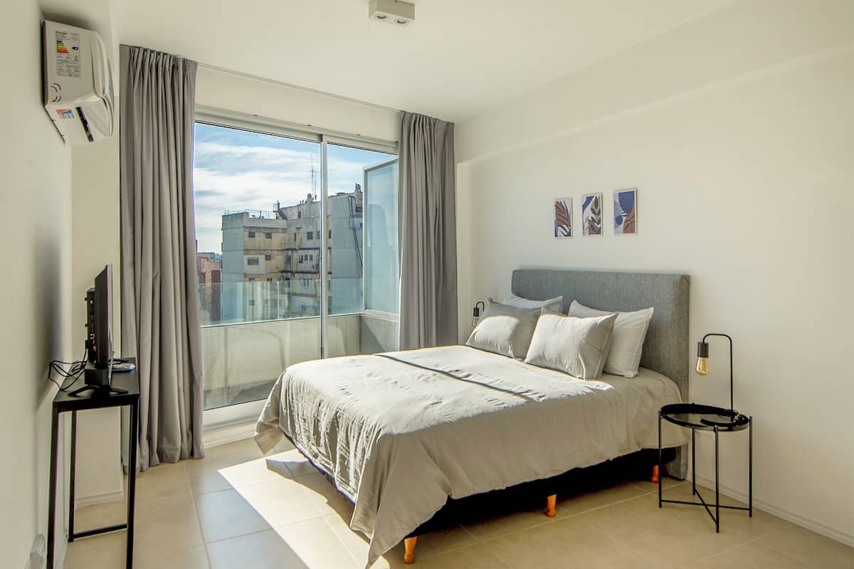 A modern bedroom with a large bed, a desk, and a view of the city through a large window.