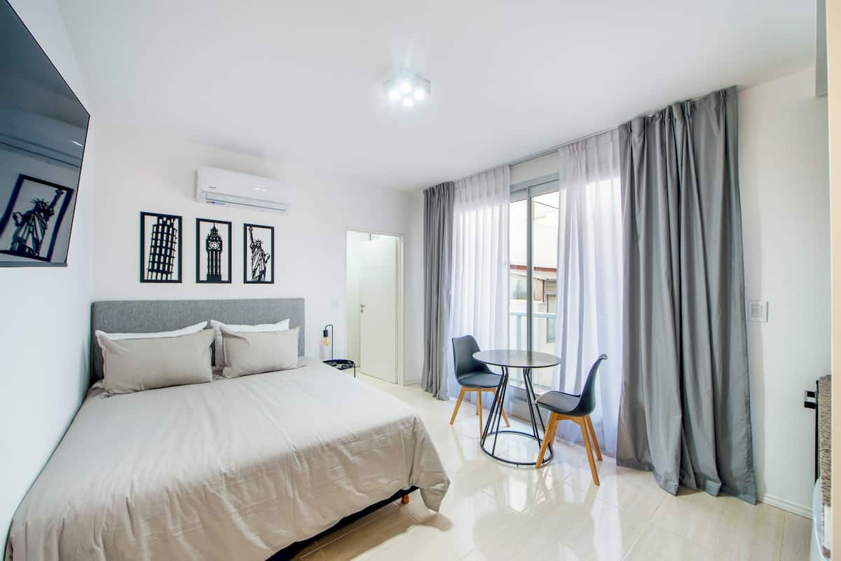 A hotel room with a bed, a small table with chairs, a window with curtains, and wall art.