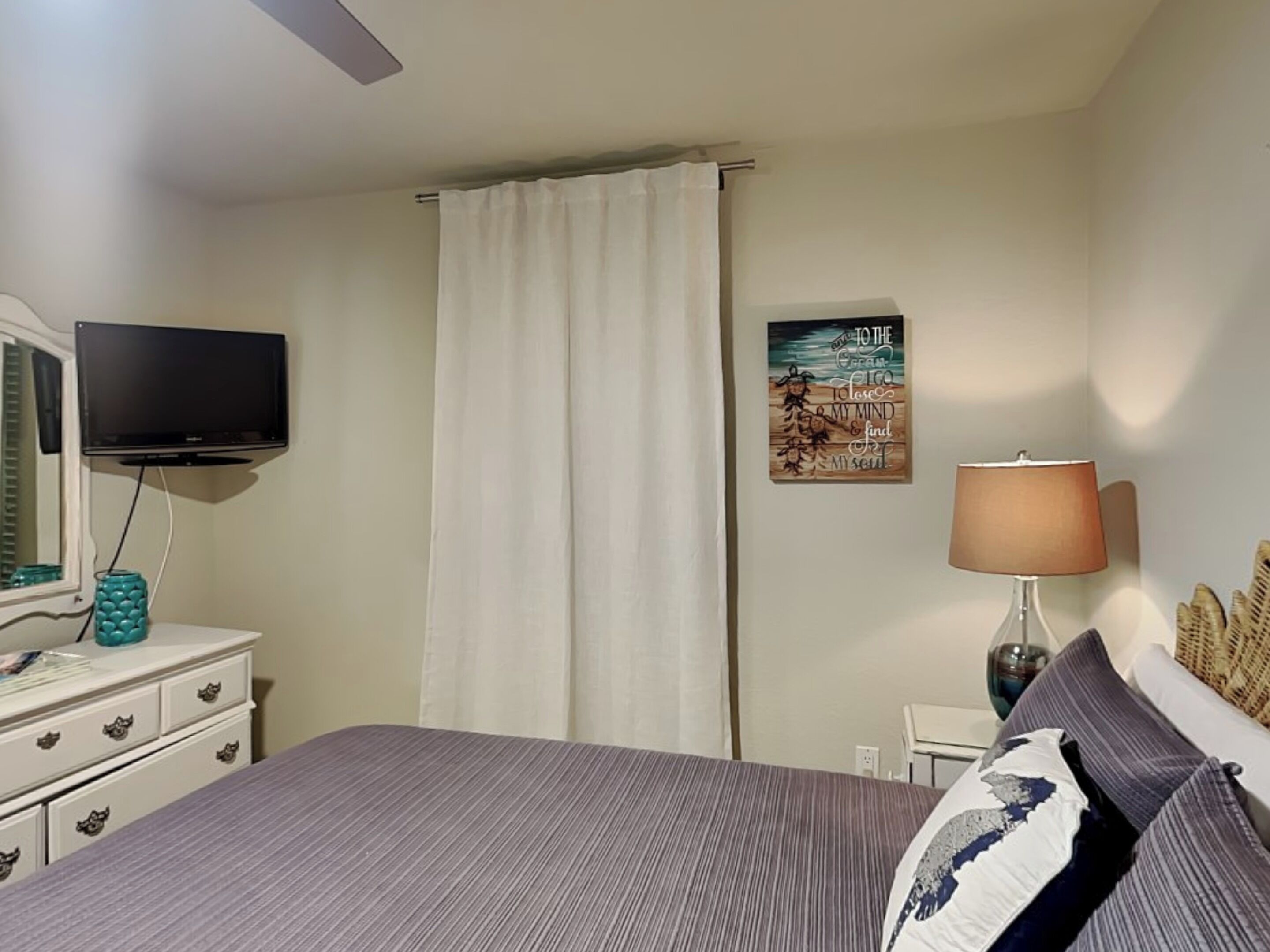 Un dormitorio con una cama, televisión, lámpara, un cuadro y una ventana con cortinas.