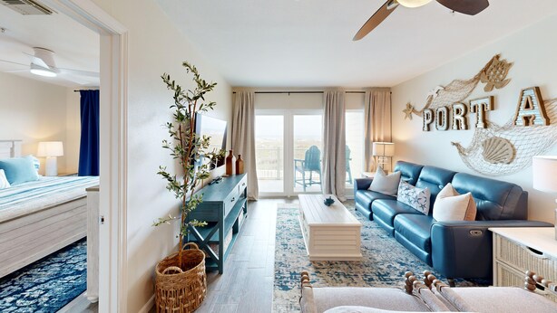 A spacious living area with a blue sofa, a wooden coffee table, and a large window with a view of the outdoors. - Grand Caribbean by Silver Sands Vacation Rentals (Port Aransas)