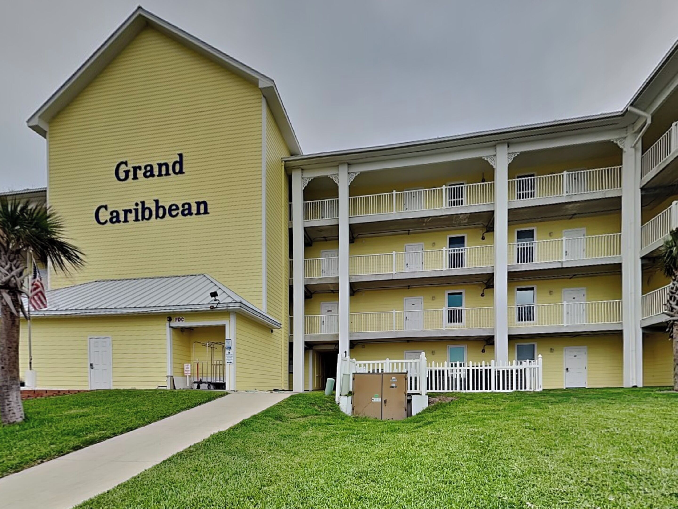 A yellow building with "Grand Caribbean" written on the side, featuring a multi-story structure with balconies and a gated entrance.