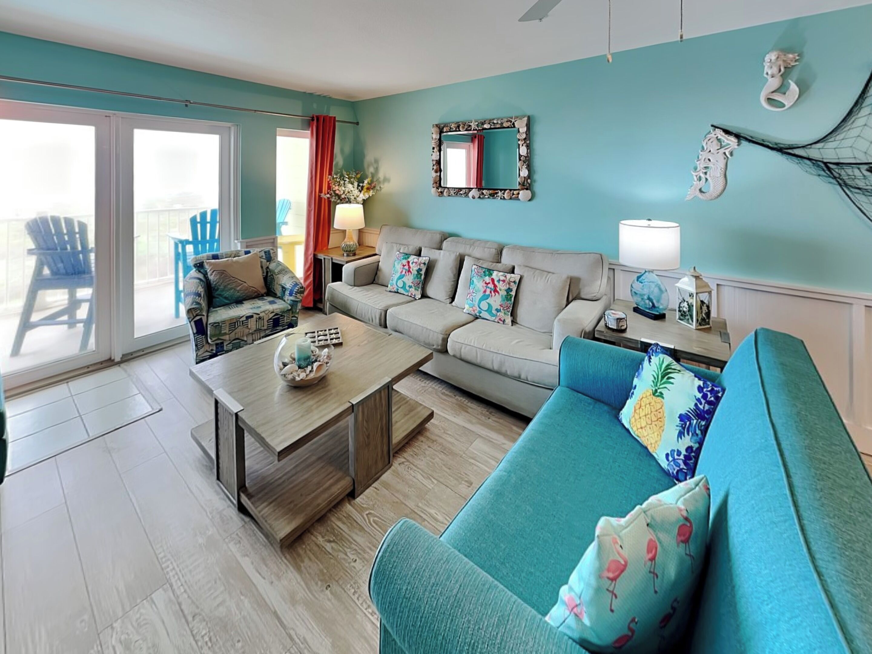 A living room with a teal sofa, a patterned armchair, a coffee table, and a sliding glass door leading to a balcony.