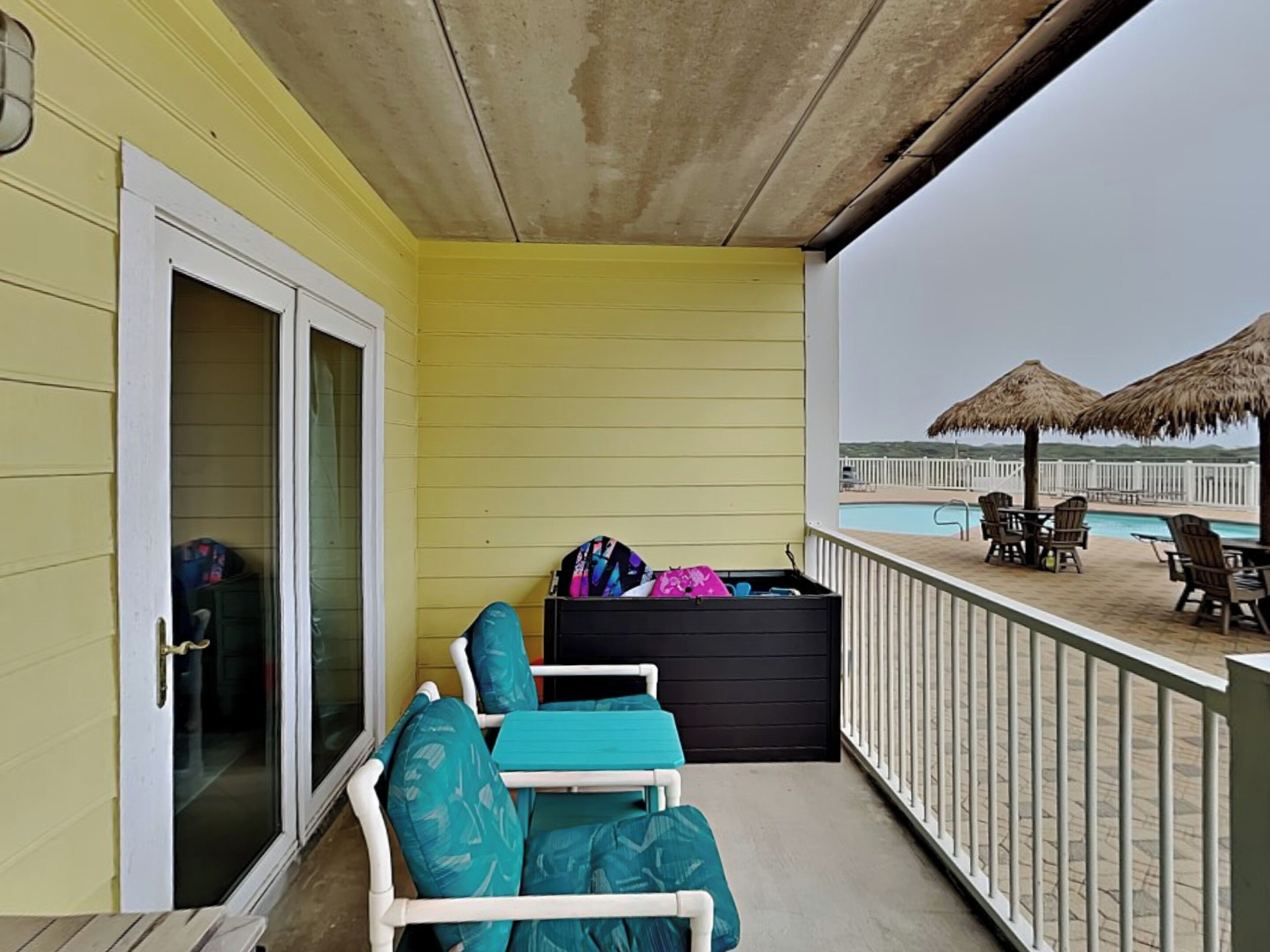 Un balcón con vista a la piscina, un mueble de almacenamiento negro y dos sillas color turquesa.