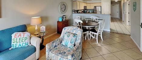 A living room with a blue sofa, patterned armchair, and a small table with a lamp. A kitchen is visible in the background with white cabinets and a dining table.