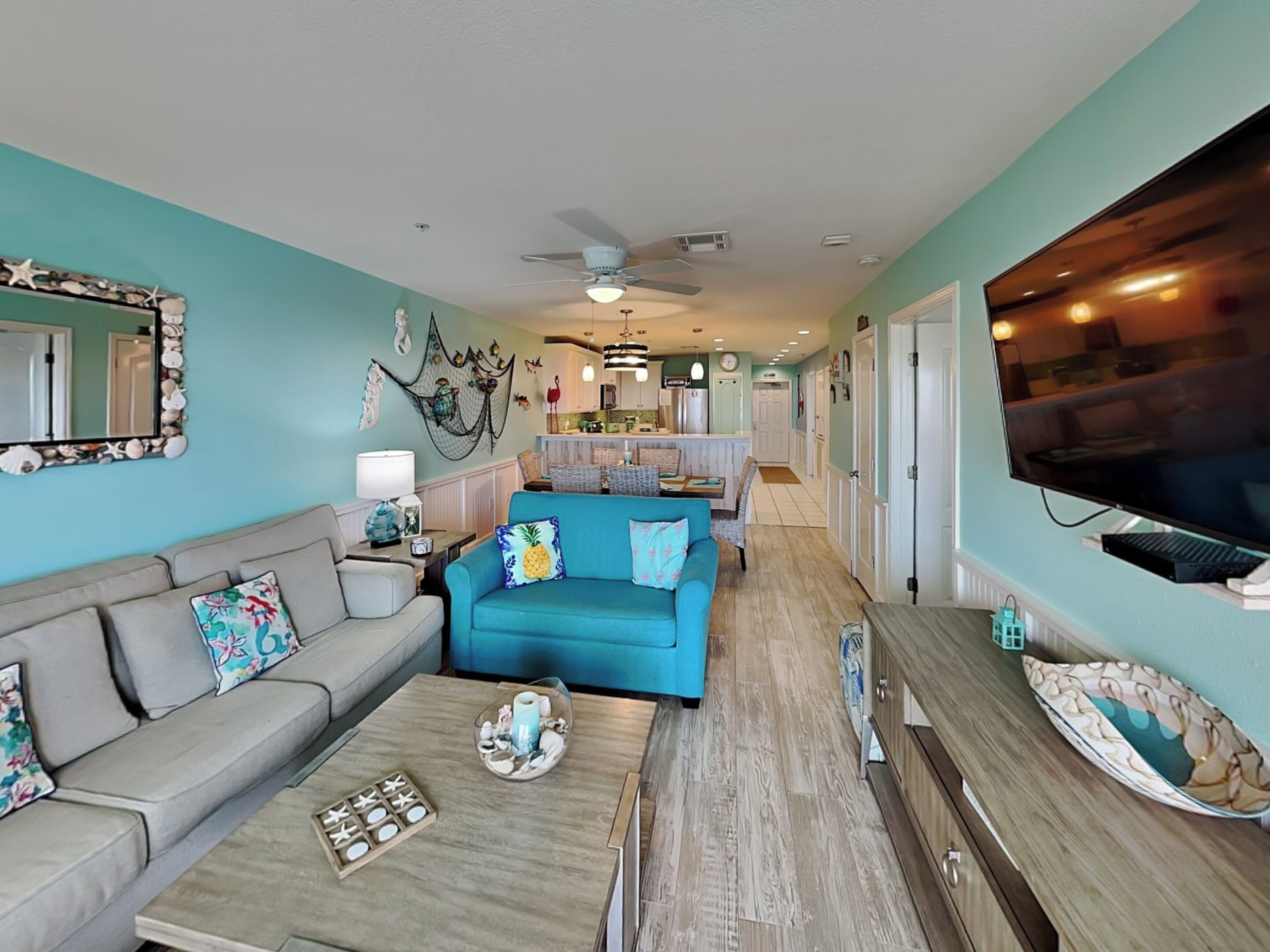 Un salon spacieux avec un canapé turquoise, un divan gris et une table basse agrémentée d’objets décoratifs. Un téléviseur est fixé au mur, et on aperçoit un coin cuisine au fond de la pièce.