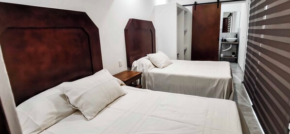 Une chambre d’hôtel avec deux lits, chacun doté d’une tête de lit en bois et de linge de lit blanc. Une petite table se trouve entre les lits, et on aperçoit une salle de bain au fond.