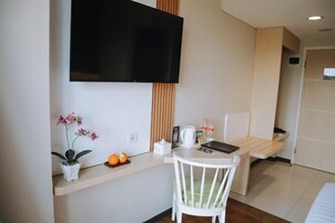 Habitación de hotel compacta con una televisión de pantalla plana montada en la pared, una mesita blanca con una silla, una planta en maceta y un frutero.