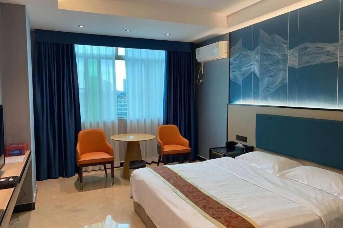 Una habitación de hotel con una cama grande, dos sillones naranja, una mesita redonda pequeña y un ventanal con cortinas.