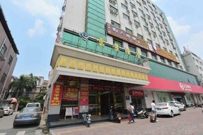 Un edificio de varios pisos con espacio comercial en la planta baja, señalización en un idioma extranjero, un toldo rojo y un estacionamiento con vehículos.