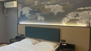 A hotel room with a bed, a nightstand, a wall-mounted air conditioner, and a decorative wall with traditional architecture and cherry blossoms. - Yunjing hotel (Dongguan)