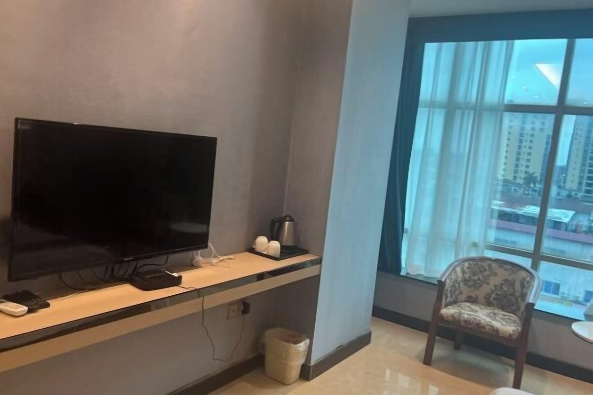 A hotel room with a large flat-screen TV on a wooden console, a coffee maker, and a chair by a large window with a city view.
