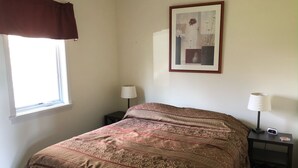 A bedroom with a patterned bedspread, a framed artwork on the wall, a bedside table with a lamp, and a window with curtains. - North Shore, Conesus Lake (Lakeville)