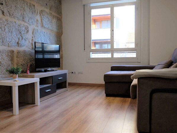 A living room with a stone wall, a flat-screen TV on a white TV stand, a brown sofa, and a window with a view of buildings. - Belvilla by OYO Villa Trabazos Abellas (Ourense)