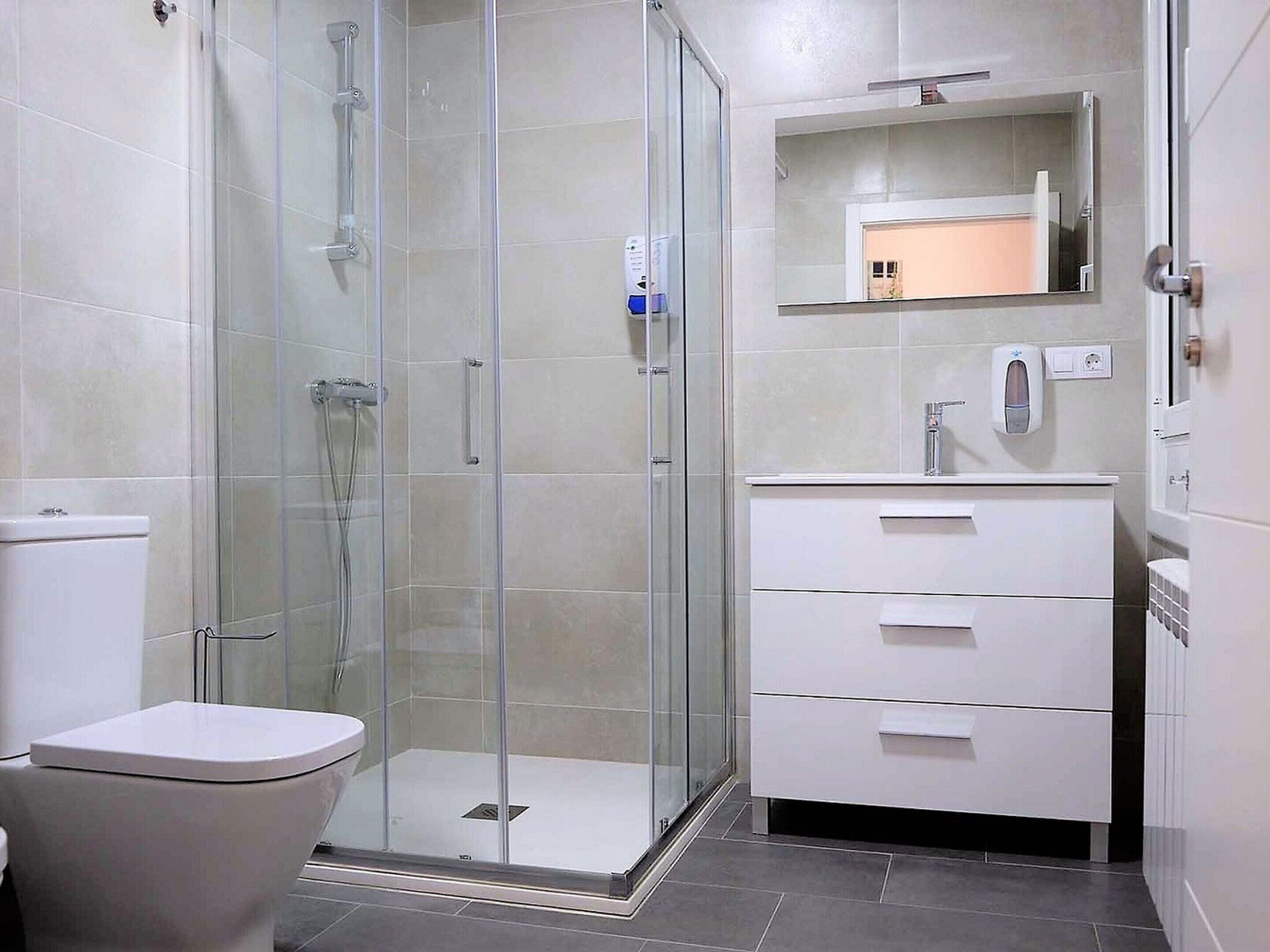 A modern bathroom with a glass-enclosed shower, a white toilet, and a vanity with a mirror and a soap dispenser.