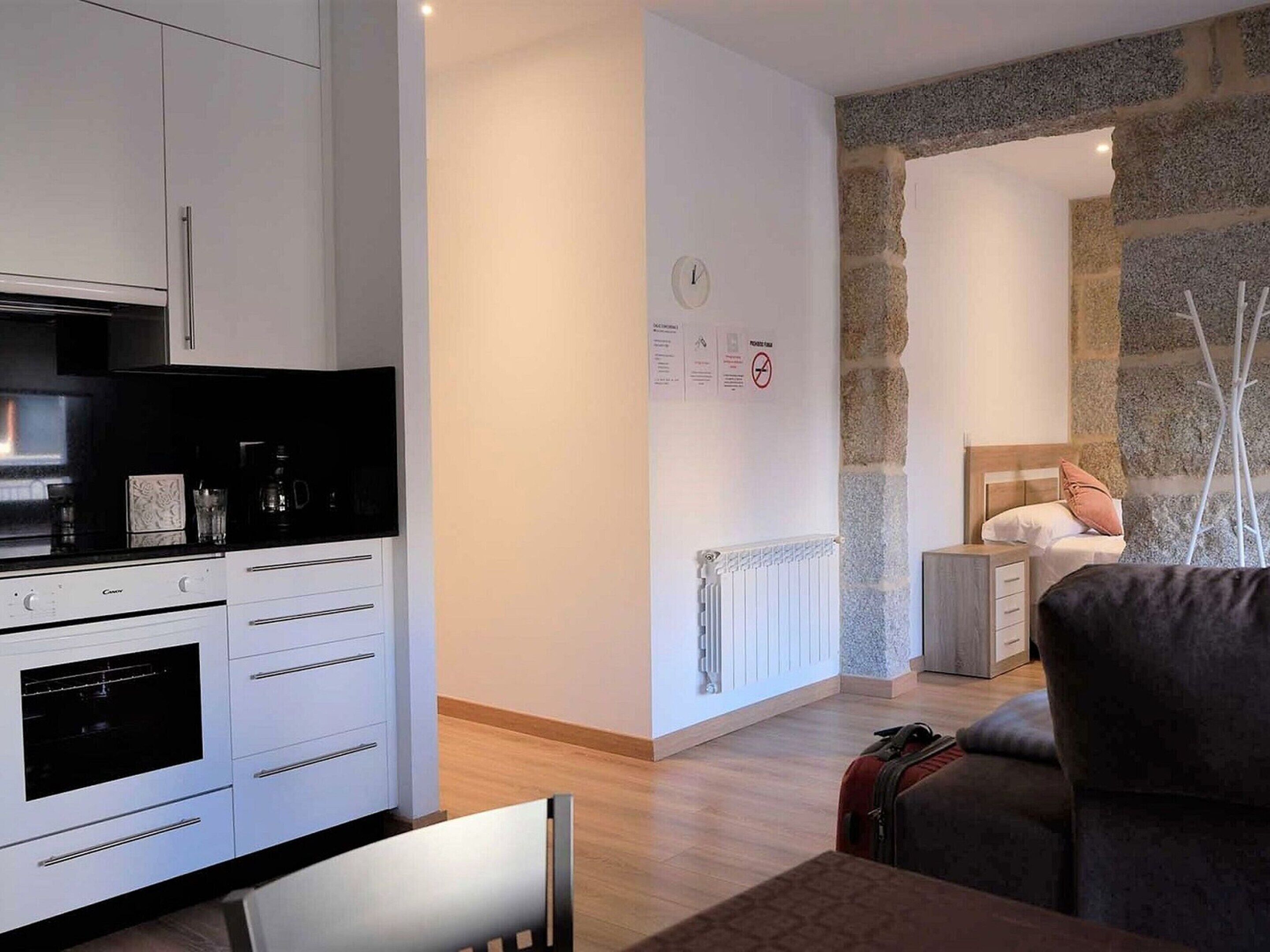 Cocina moderna con armarios blancos, encimera negra y horno empotrado. En la habitación contigua, una pared de piedra, un radiador y una cama con cabecero de madera.
