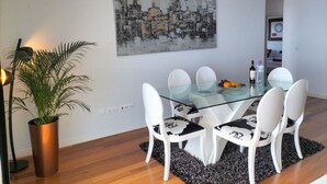 A dining area with a glass table, white chairs, a potted plant, and a black and white cityscape painting on the wall.