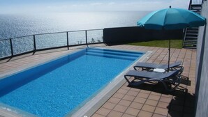A rectangular swimming pool with clear blue water, a tiled deck, and a blue umbrella providing shade.