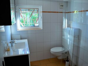 Ein Badezimmer mit weißem Waschbecken, Toilette und einem Fenster mit Blick ins Grüne.