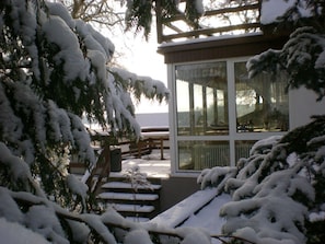 Ein mit Schnee bedeckter Balkon mit einer hölzernen Bank und einem Tisch, der zu einem Glastür-Eingang eines Gebäudes führt.