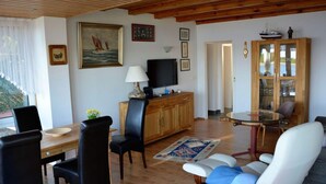 A cozy living room with a wooden ceiling, a dining area with a table and chairs, a television, and a cabinet with glass doors. - Holiday House House by the Lake (Krakow am See)