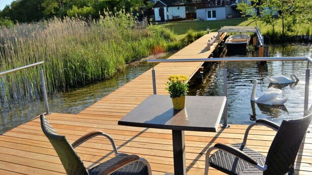 A wooden deck with a table and chairs overlooking a calm body of water, with a house and greenery in the background. - Holiday House House by the Lake (Krakow am See)