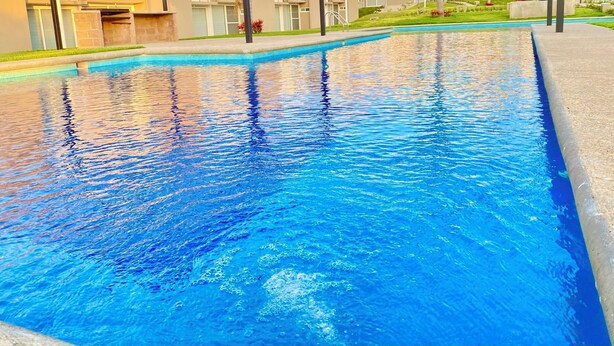 A swimming pool with clear blue water, surrounded by a concrete deck and a grassy area with a building in the background. - Alojamiento en Yecapixtla, Morelos. Disfruta con Amigos o Familia! (Yecapixtla)