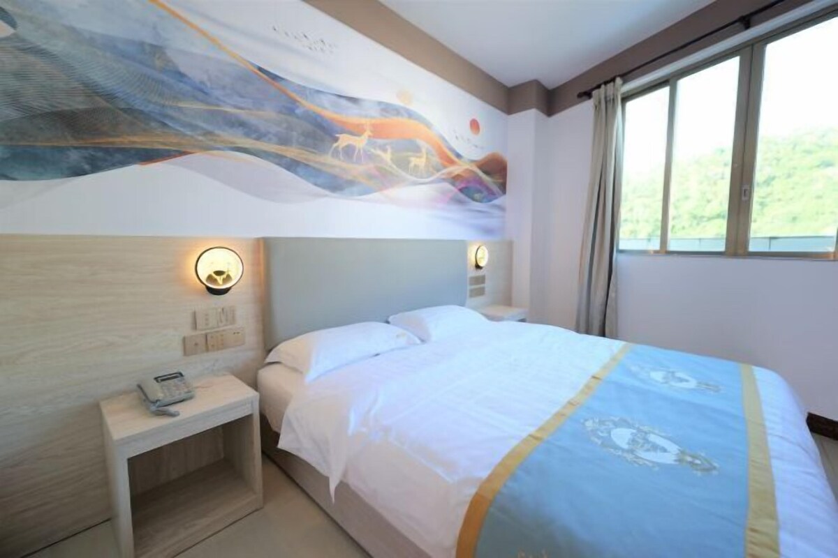 Una habitación de hotel con cama, lámparas de noche, mesita de noche con teléfono y un ventanal con cortinas.