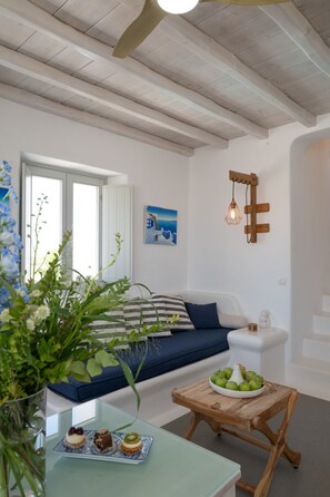 Una habitación con una cama, una mesita con frutero, una lámpara de pared y una ventana con contraventanas blancas.