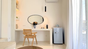 Un baño moderno con un lavamanos blanco, un espejo redondo, una silla de madera, un refrigerador pequeño y un aire acondicionado de pared.