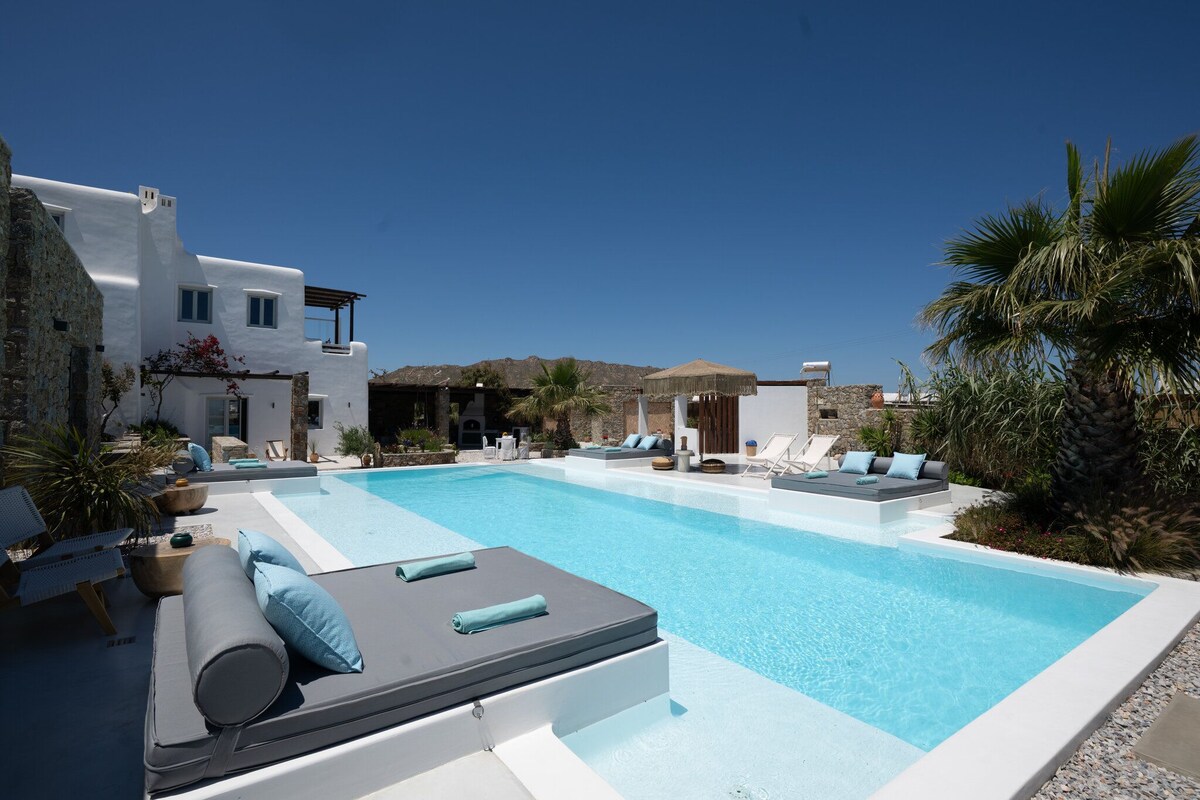 Un espace piscine avec des chaises longues et un bâtiment blanc agrémenté d’éléments en pierre.