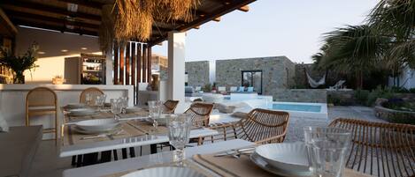 An outdoor dining area with tables set for a meal, a pool, and a stone wall in the background.