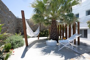 A white building with a stone wall, a hammock, a palm tree, and a white lounge chair. - Myconian Theros (Mykonos)