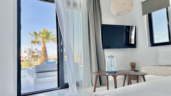 Una habitación moderna con un amplio ventanal corredero que da a un área de piscina exterior, un televisor de pantalla plana montado en la pared, una pequeña mesa redonda con un recipiente de vidrio y una zona de estar acogedora con un sofá y una silla.